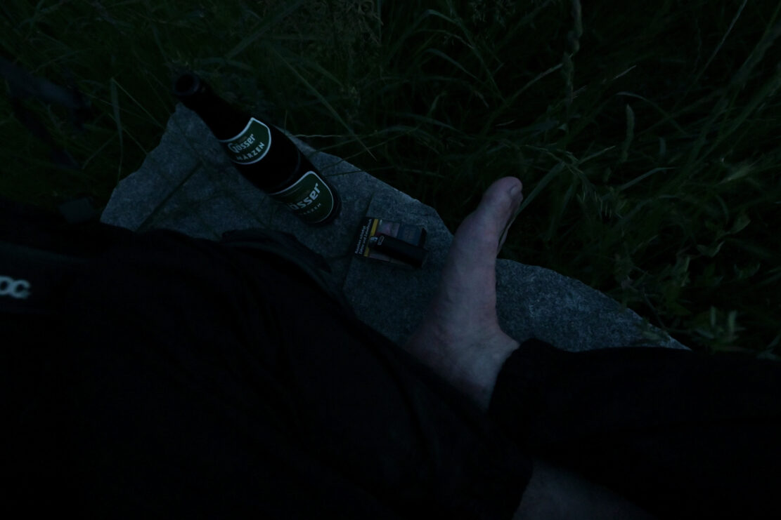 Füße eines Mannes, der im Schneidersitz auf einem Stein sitzt, mit Zigarretten und Bier neben sich im letzten Abendlicht.