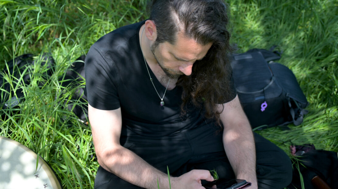 Alter Mann mit langen Haaren sitzt in der Wiese und schaut aufs Handy.
