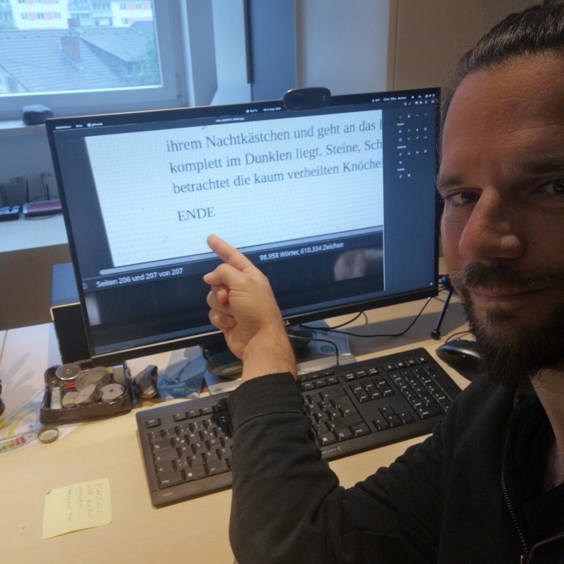 James zeigt auf einen Monitor, auf dem das Wort ENDE seines Romans zu lesen ist.