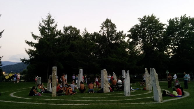Das Drum & Dance im Steinkreis im Klagenfurter Europapark. Menschen stehen und sitzen zusammen in einem modernen Steinkreis und machen zusammen Musik. Einige tanzen.